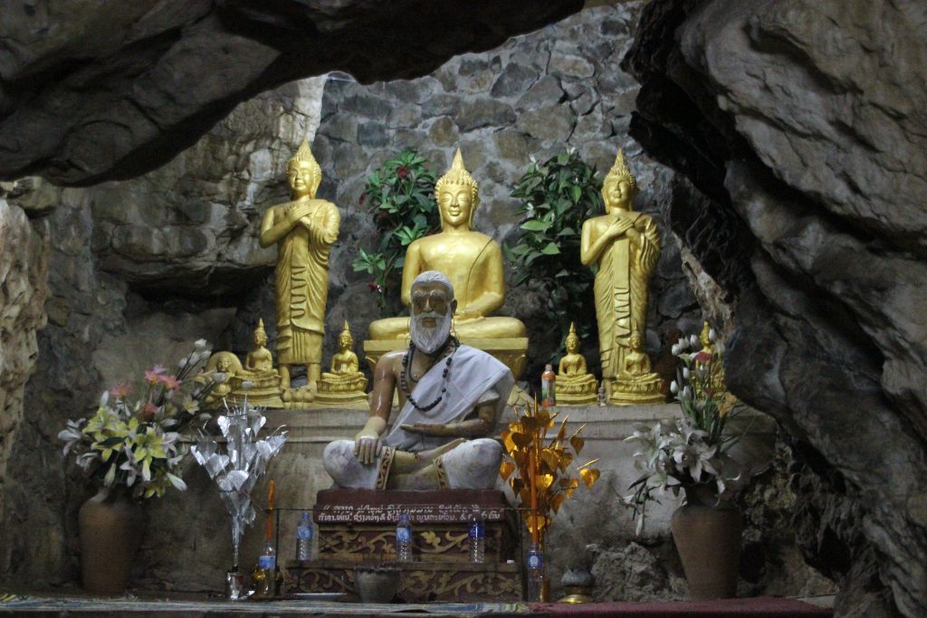 Mönchsstatue vor goldenen Buddhafiguren in einem Höhlentempel